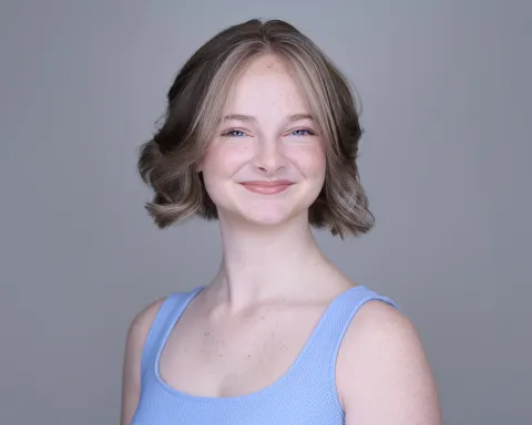 Young actor with wavy bob hairstyle smiling warmly in light blue tank top against gray backdrop
